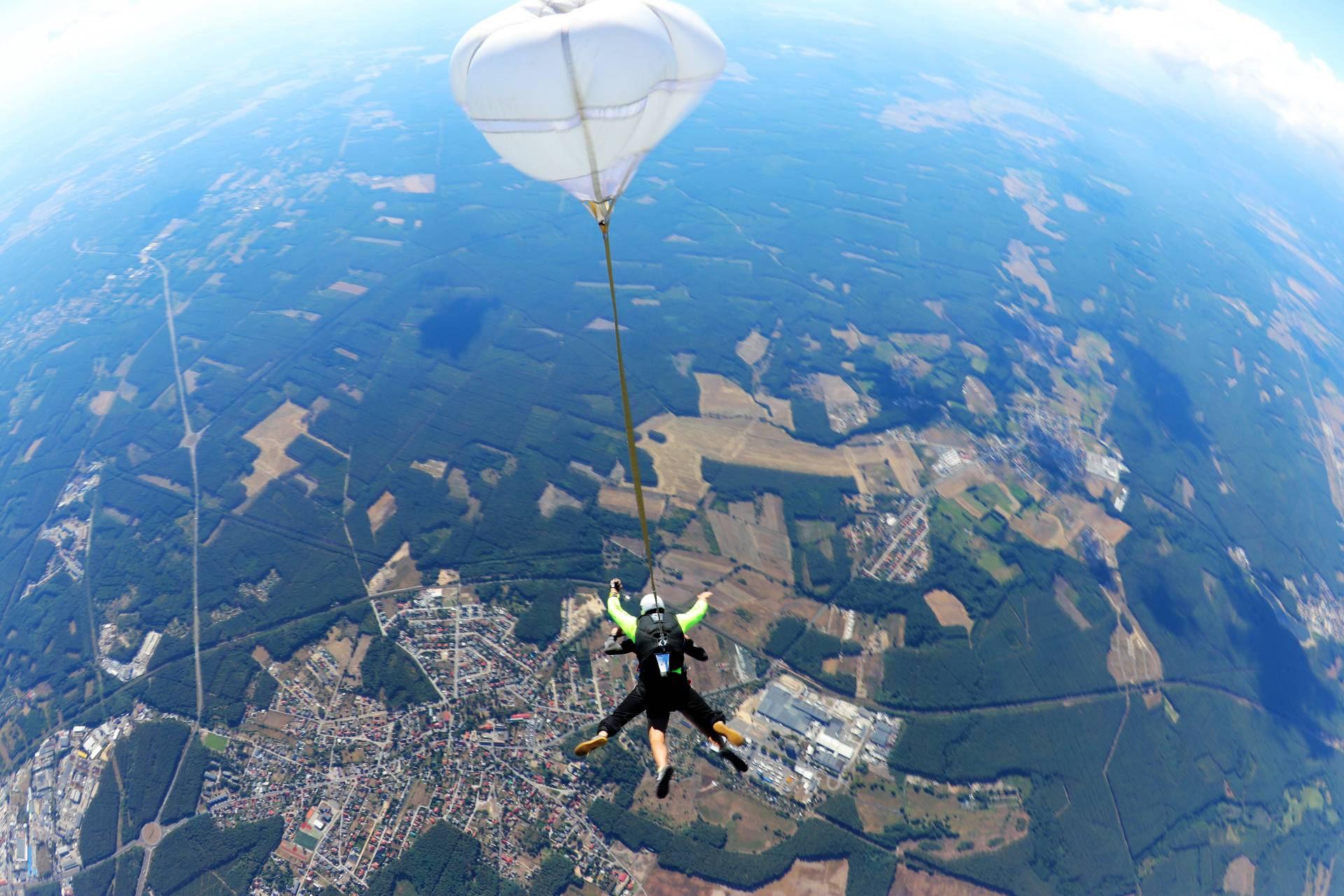 Tandem ze spadochronem hamującym nad polami i zabudową.